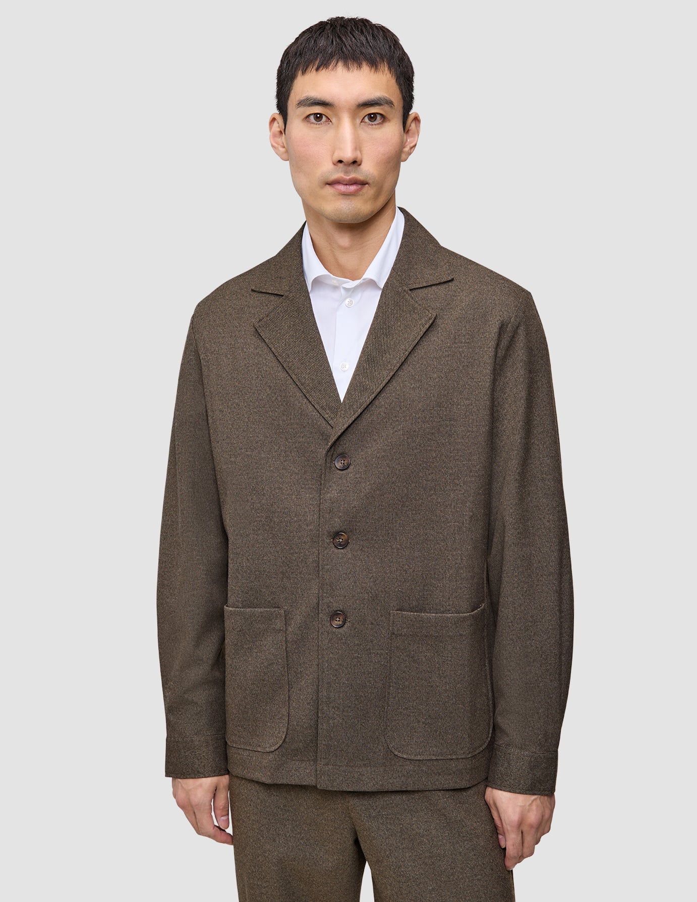 En man klädd i en mörkbeige Atelier Overshirt i brutet twill över en vit skjorta med krage.
